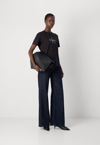 T-shirt noir en coton avec logo, jeans évasés bleu foncé, et une pochette en cuir noir structurée. Mannequin portant des talons aiguilles noirs.