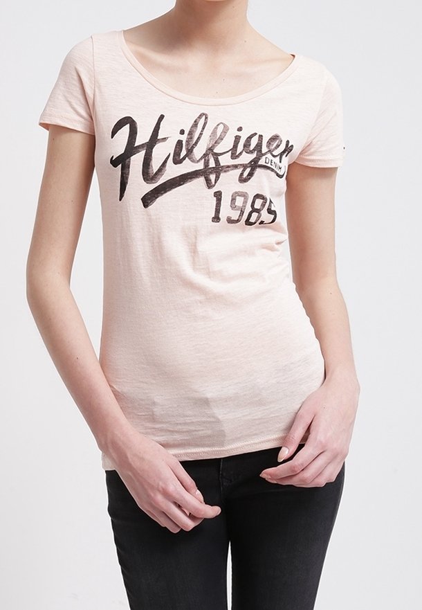 Světle růžové tričko s krátkým rukávem a kulatým výstřihem, na kterém je černý potisk loga a rok "1985" na prsou. Hladká textura.