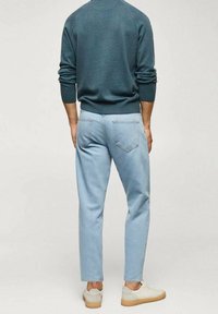 Hellblaue Jeans mit geradem Schnitt, getragen zu einem dunkelgrünen Pullover; kombiniert mit weißen Schuhen mit brauner Sohle.