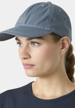 Mujer joven de piel clara, ojos verdes y cabello trenzado que lleva una gorra de béisbol azul y una camiseta azul oscuro, mirando ligeramente hacia la izquierda.