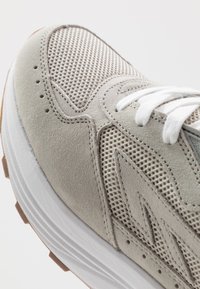 Lys grå sneaker med suede og mesh-materialer. Distinktiv tekstureret overflade og hvide snørebånd med en kontrasterende hvid sål.