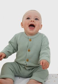 Grüner gerippter Strick-Babyanzug mit Holzknöpfen, langen Ärmeln und Bündchenhosen, die auf einer weißen strukturierten Oberfläche sitzt.