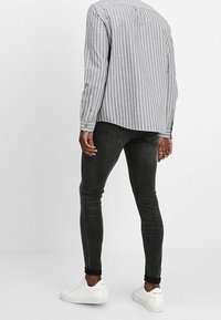 Jeans neri aderenti con sfumature sottili, abbinati a una camicia a righe grigio chiaro e bianca con bottoni. Presenta sneaker bianche classiche.
