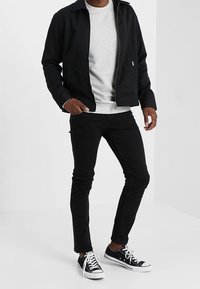 Chaqueta negra con cremallera sobre un sudadera gris, combinada con jeans negros ajustados y zapatillas negras con suelas y cordones blancos.