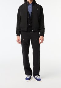 Homme portant une veste noire, un pantalon noir, un polo bleu et des baskets bleues avec des lacets blancs sur fond blanc.