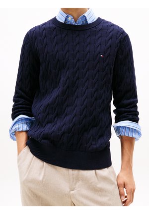 Homme portant un pull en maille torsadée bleu marine par-dessus une chemise à rayures bleues avec manches retroussées et un pantalon beige, mains dans les poches.