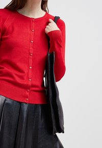 Woman wearing a red buttoned cardigan and black leather skirt, holding a large black tote bag over her shoulder.