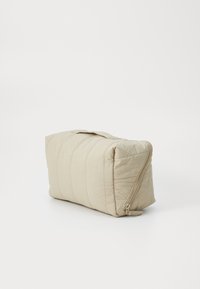 Beige, kviltet kosmetikkveske med rektangulær form, teksturert overflate og glidelås, med et stoffhåndtak på toppen.