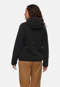 Veste en polaire noire avec capuche, tissu texturé, coupe décontractée et un détail de logo au dos. Associée à un pantalon beige.