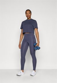 Jūras spēku stila krūšturis, pārī ar saskaņotiem augsta vidukļa legingiem, abi izgatavoti no elastīga materiāla. Modelis tur zilus hanteles, valkājot baltas sporta kurpes.