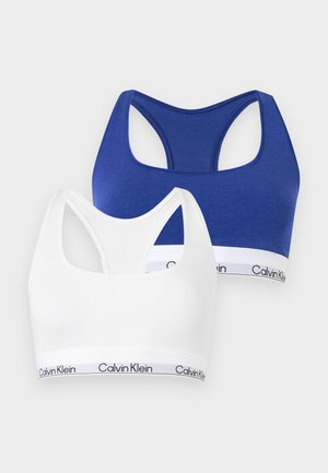 Młoda kobieta stoi boso, ubrana w biały zestaw bielizny marki Calvin Klein na jednolitym jasnym tle.