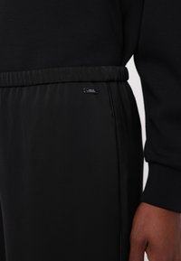 Fekete sportos joggerek, puha tapintású anyaggal, elasztikus derékkal és egy kis fém márkacímkével az oldalon. Közeli nézet az anyagról és a részletekről.