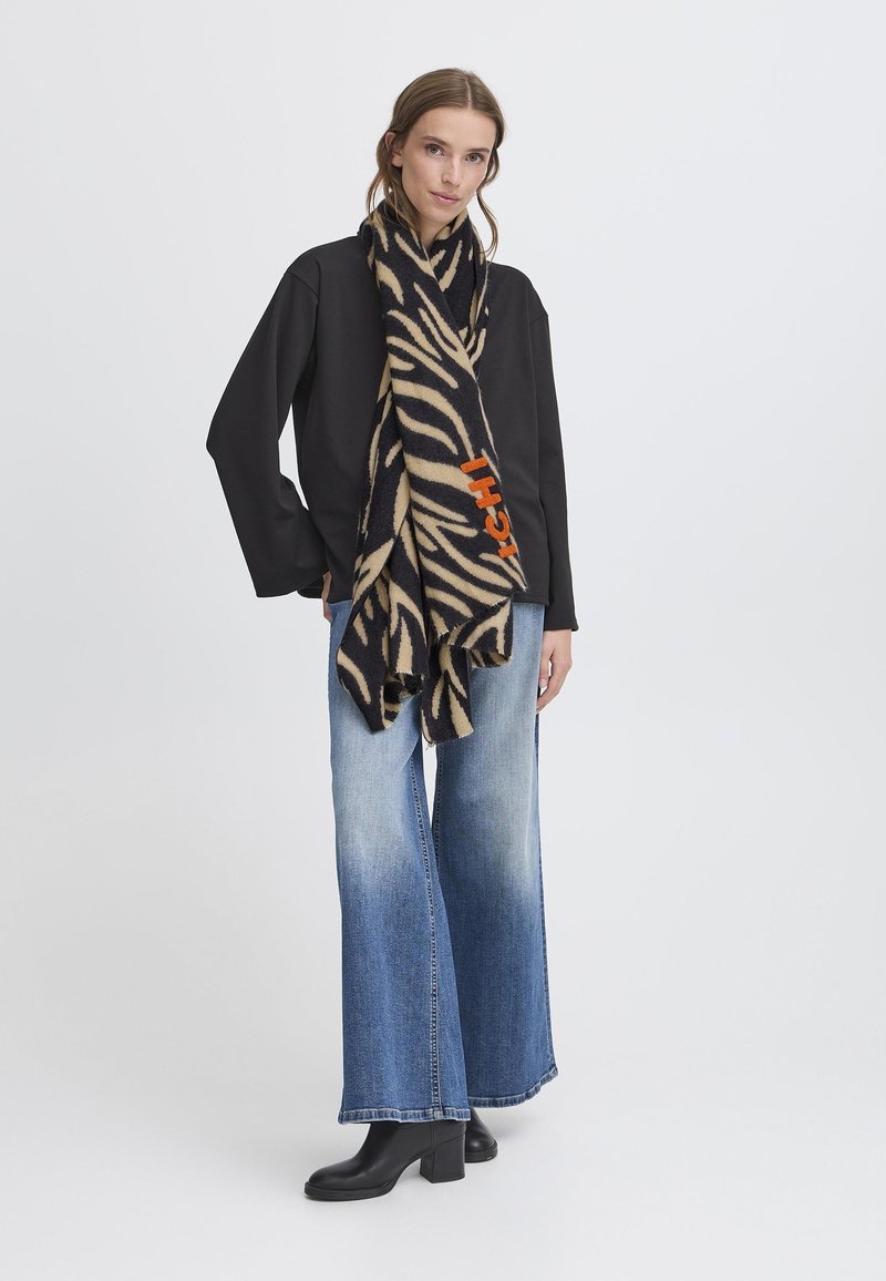 Sciarpa a motivo zebra nera e beige realizzata in morbida lana, con un logo arancione. Indossata con una maglietta nera e jeans a campana blu.
