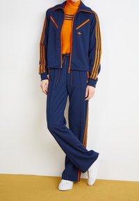 Mörkblå träningsdräkt med orange detaljer, bestående av en zip-up jacka och byxor med dragsko. Vita sneakers fulländar outfiten.