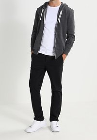 Felpa grigia con zip e cordini, t-shirt bianca sotto, pantaloni neri e sneakers bianche; design semplice e vestibilità casual.