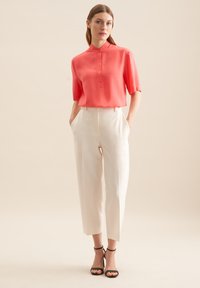 Blusa corallo a maniche corte con colletto alto e patta, abbinata a pantaloni bianchi su misura. Tacchi neri con cinturino alla caviglia completano l'outfit.