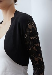Svart kortärmad cardigan med långa ärmar i spets. Kroppen är i solid svart stickad kvalitet, medan ärmarna har blommig spetsdetalj.