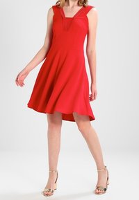 Robe rouge sans manches avec un corsage ajusté, un décolleté en V et une jupe évasée. Présente un tissu texturé et un ourlet haut-bas. Associée à des talons métalliques.