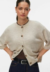 Bež kratka jopica s kratkimi rokavi z zaobljenim ovratnikom, z petimi rjavimi gumbi in ohlapnim prileganjem. Oblečena čez temno rjavo majico in denim.