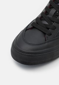 Baskets en cuir noir avec semelle épaisse texturée et lacets noirs, coutures détaillées et accents rouges subtils près de la languette.
