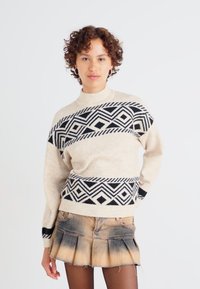 Beige gebreide trui met een zwart geometrisch patroon, voorzien van een hoge hals en lange mouwen. Gecombineerd met een denim minirok.