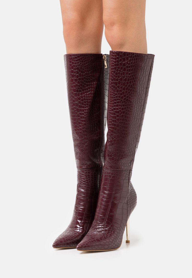 Burgundy saapaat, jotka ulottuvat polveen, ovat valmistettu kiiltävästä matelijanaamioidusta materiaalista. Niissä on terävä kärki ja kapea stiletto-korko, sekä sivuvetoketju.