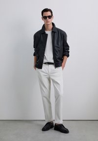 Uomo con giacca di pelle nera, camicia bianca, pantaloni bianchi, cintura nera, scarpe nere e occhiali da sole neri, in piedi contro una parete grigia uniforme.