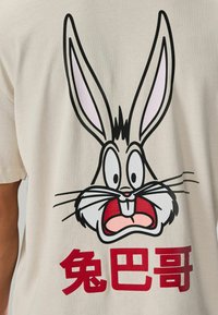 Hellbeige T-Shirt mit einem großen, comicartigen grauen Hasenkopf-Design mit rosafarbenen Ohren und rotem Text darunter.