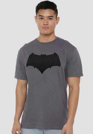 Young man wearing a gray t-shirt with a large black Batman logo on the chest, standing against a plain background.