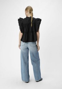 Schwarze, gestufte Baumwollbluse mit gezackten Rüschen an den Ärmeln, kombiniert mit hochgeschnittenen, hellblauen, weiten Jeans. Einfarbiger Hintergrund.