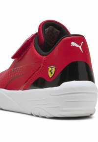 Czerwono-czarny sneaker z gładką syntetyczną cholewką, wyposażony w pasek na rzep, logo Puma i emblemat Ferrari, na białej gumowej podeszwie.