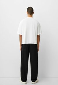 Bílá tričko s krátkým rukávem a volným střihem, spárované s černými širokými kalhotami a světlými teniskami, pohled zezadu.