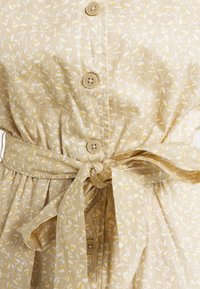 Knapplång klänning i ljusbeige bomull, med ett blommönster i vitt och gult. Täljdes vid midjan med ett matchande bälte och flera knappar.