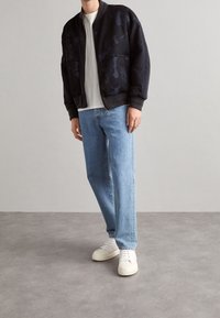 Person står i avslappnad klädsel: en svart kamouflagemönstrad bomberjacka, vit t-shirt, blå jeans och vita sneakers, mot en neutral bakgrund.