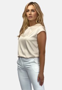 Jasnobeżowa bluzka z krótkim rękawem, okrągłym dekoltem i miękką teksturą, w zestawieniu z dopasowanymi białymi spodniami. Czysty i minimalistyczny design.