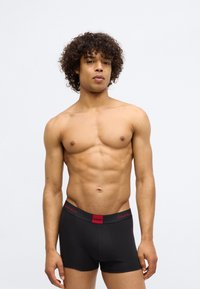 Boxers pretos com a cintura vermelha com o logótipo "Calvin Klein", design justo e um tecido suave e elástico, exibidos em um modelo masculino sem camisa.