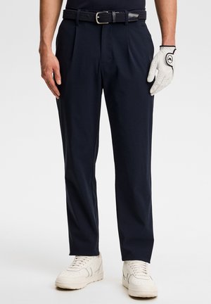 Mann trägt dunkelblaue, plissierte Hose mit gewebtem Gürtel, weißen Golfhandschuh an der rechten Hand und weiße Sneaker vor weißem Hintergrund.