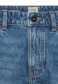 Close-up van de tailleband van een blauwe spijkerbroek met knoopsluiting, lussen voor een riem en het merklabel "camel active" aan de binnenzijde van de tailleband.