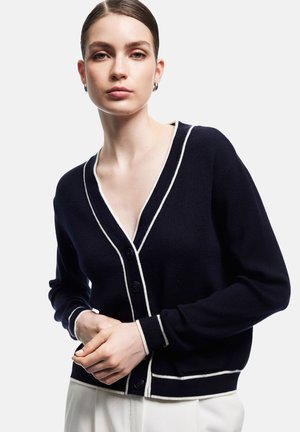 Granatowy kardigan z białym wykończeniem, dekoltem w serek, długimi rękawami i zapięciem na guziki. Dzianinowy materiał charakteryzuje się gładką fakturą i swobodnym krojem.