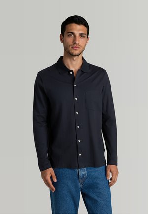 Junger Mann mit kurzen dunklen Haaren, der ein schwarzes Hemd mit langen Ärmeln und blaue Jeans vor einem einfachen hellgrauen Hintergrund trägt.