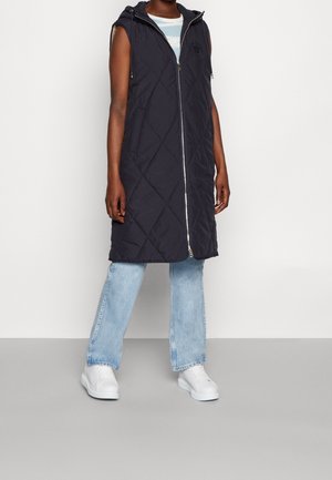 Personne portant une longue veste noire matelassée sans manches par-dessus une chemise rayée, un jean bleu clair et des baskets blanches, debout contre un mur uni.