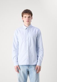 Chemise boutonnée rayée bleu clair avec un col, des manches longues et un petit logo sur la poitrine. Portée avec un jean bleu clair.
