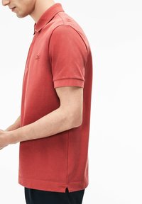 Röd polotröja i bomull, med krage, korta ärmar och en liten logotyp. Botten har sidoflikar för ökad rörelsefrihet.