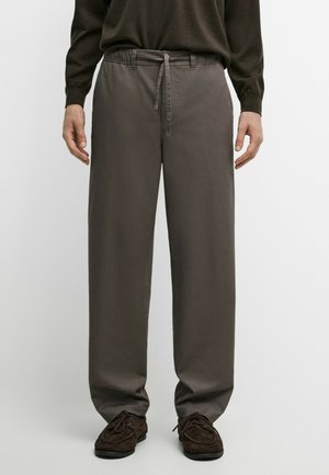 Person trägt lockere graue Hose mit Kordelzug in der Taille, dunkelbraune Schuhe und ein dunkles Langarmoberteil vor einfarbigem Hintergrund.