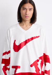 Hvid mesh jersey med stort rødt Nike-logo og grafiske detaljer. V-hals design med lange ærmer og tekstureret mønster over det hele.