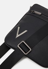 Černá crossbody kabelka z umělé kůže s texturovaným povrchem, předním kapsou na zip, stříbrným kováním a výrazným stříbrným logem ve tvaru "V".