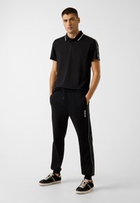 Czarny polo z białym kołnierzykiem, w zestawie z czarnymi spodniami dresowymi z detalami logo i białymi paskami po bokach. Uzupełnione czarnymi trampkami.