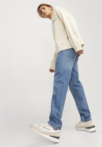 Bež majica dugih rukava s izvezenim tekstom, svijetloplave traperice ravnog kroja i kremasto-bele tenisice s tamnim detaljima, nošeni s kačketom.