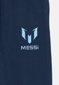 Tumšzilas sporta bikses ar gaiši zilu logo, kurā ir stilizēts 'M' un uzraksts 'MESSI', kas drukāts uz kreisā kājas. Izgatavotas no mīksta auduma.