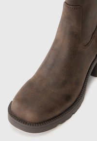 Hnědá kožená kotníková bota s kulatou špičkou, texturovaným povrchem a žebrovanou podrážkou. Nabízí robustní design se subtilními švy.